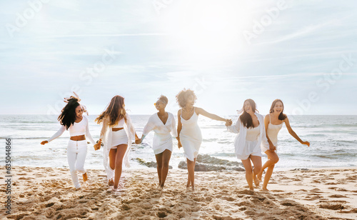 Group of joyful diverse women enjoying hen party, running and having fun on the beach at coastline, full length