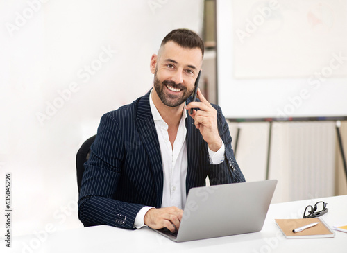 Smiling caucasian mature man with beard in suit sit at table, use laptop, making phone calls