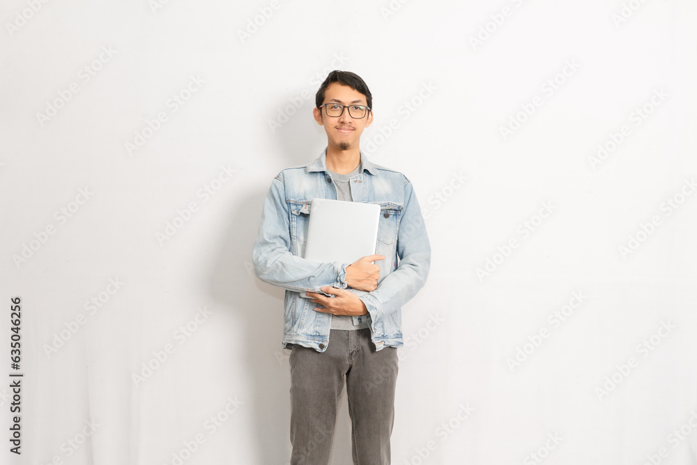 A male gives happy expression with confident while holding his laptop. Indonesian or southeast asian model isolated with white wall background. A male gives happy expression with confident while holding his laptop. Indonesian or southeast asian model isolated with white wall background.