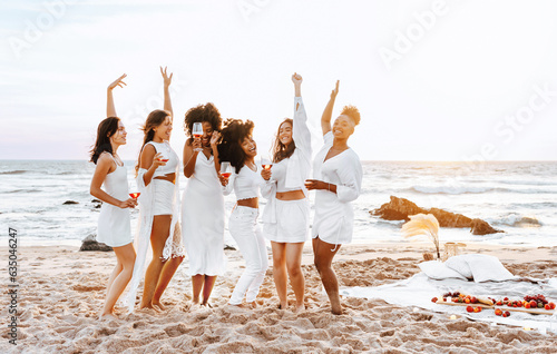 Hen party picnic. Young diverse ladies with glasses of wine dancing standing on the beach in the evening photo