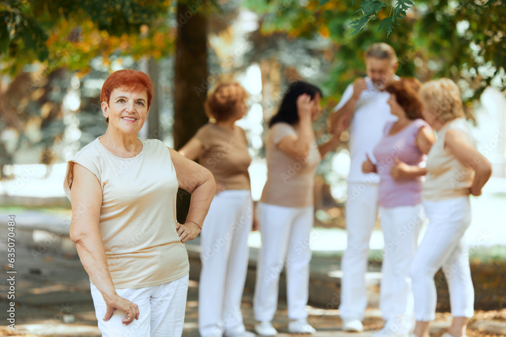 Focus on senior redhead smiling woman standing in park on warm sunny day. Blurred people on background. Concept of sport and health, active lifestyle, age, wellness, body, care