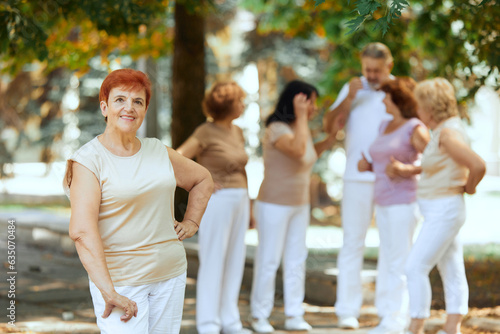 Focus on senior redhead smiling woman standing in park on warm sunny day. Blurred people on background. Concept of sport and health  active lifestyle  age  wellness  body  care