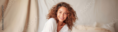 Portrait of a fictional happy, smiling beautiful woman with naturally curly auburn colored hairs on a blurred soft, textured background with pillows. Concept of happiness, good sleep and feel-good.  