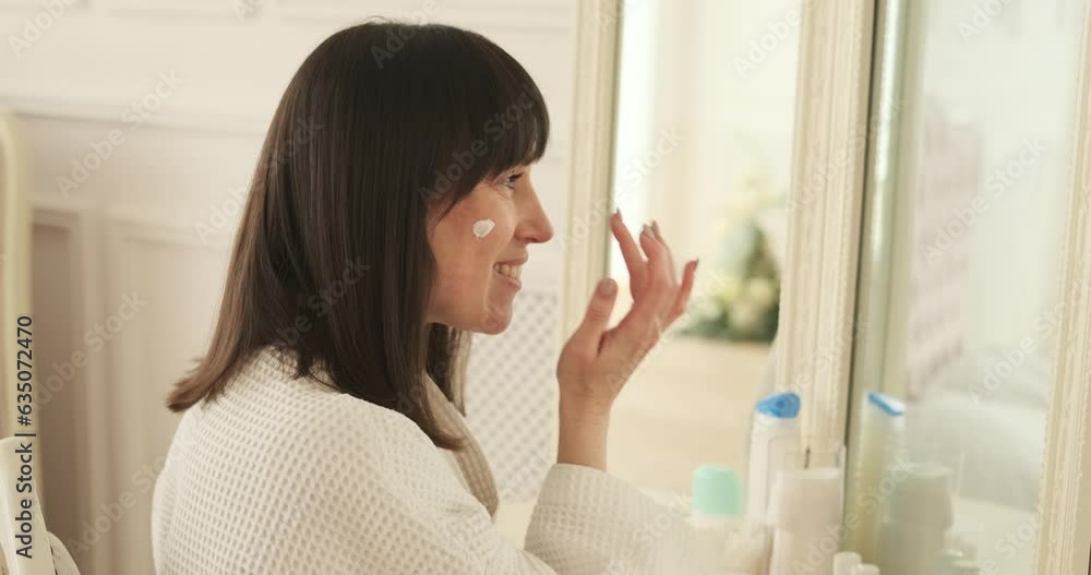 Content woman is seen using facial lotion near the vanity. Soft lighting accentuates the tranquil ambiance, creating a comfortable and soothing space for her skincare routine. Content woman is seen using facial lotion near the vanity. Soft lighting accentuates the tranquil ambiance, creating a comfortable and soothing space for her skincare routine.