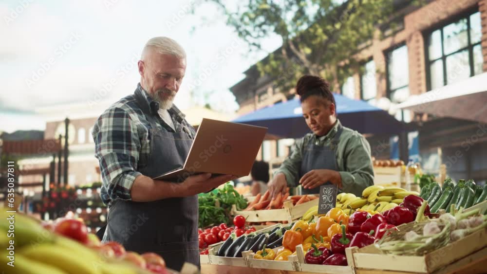 Market Manager Using Laptop Computer while Standing Next to Street Vendor Stalls with Fresh Organic Agricultural Products. Adult Male Managing Business Development in Rural Farmers Community Market Manager Using Laptop Computer while Standing Next to Street Vendor Stalls with Fresh Organic Agricultural Products. Adult Male Managing Business Development in Rural Farmers Community