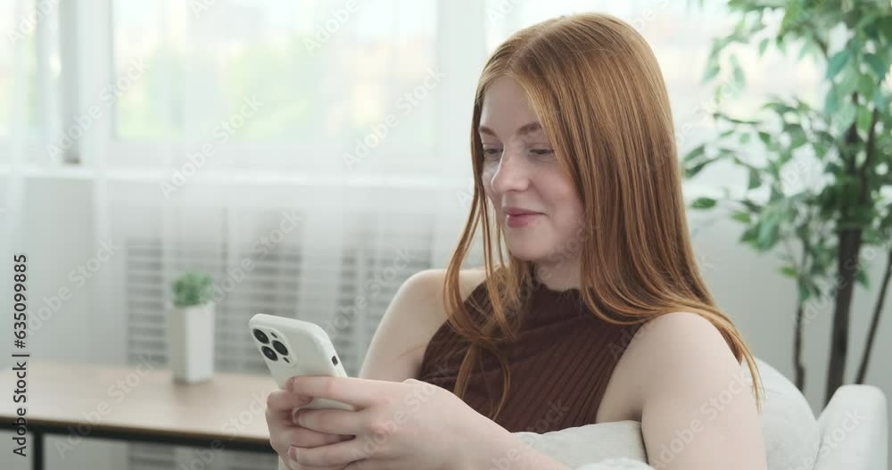 Cheerful red haired teenage girl is captured texting on her phone while sitting on a cozy sofa. Her genuine smile fills the room with positivity, and her fingers dance across the phone screen. Cheerful red haired teenage girl is captured texting on her phone while sitting on a cozy sofa. Her genuine smile fills the room with positivity, and her fingers dance across the phone screen.