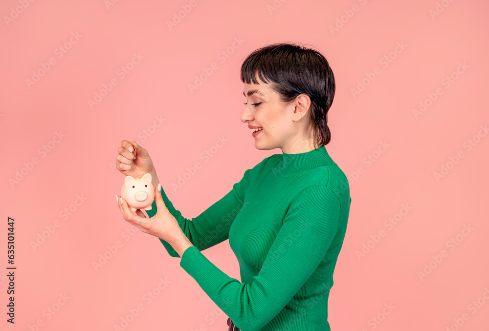 Joyful woman wearing green casual sweater saving money for rainy day in the piggy bank, investment, saving money, currency, deposit. Indoor studio shot isolated on pink background.