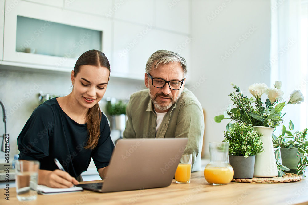 A smiling woman writes notes while using a laptop with her father at home. A smiling woman writes notes while using a laptop with her father at home.