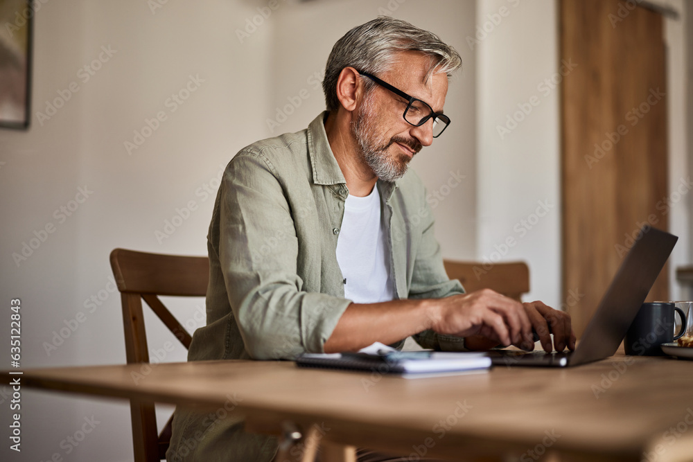 A mature man in casual clothing sitting at the table and working on a laptop.