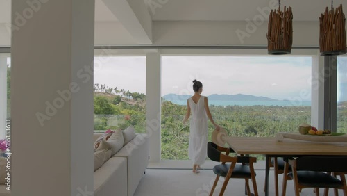 Young woman in white summer dress with hat in hand admires wonderful view of jungles glass on open terrace of spa resort room, tropical holiday concept