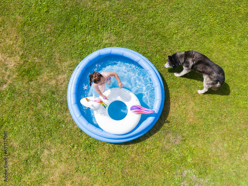 A little girl splashes in an inflatable pool in the garden, a husky dog ​​stands nearby.  Drone photo.