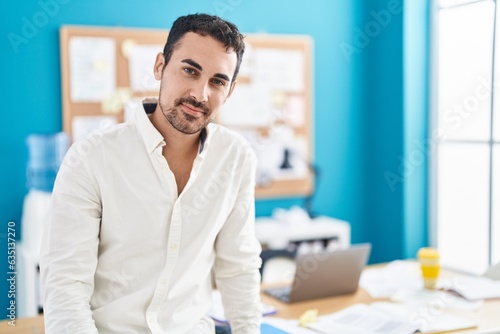 Young hispanic man business worker smiling confident sitting on table at office