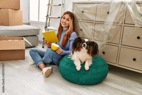 Young caucasian woman drinking coffee reading book sitting on floor with animal at new home