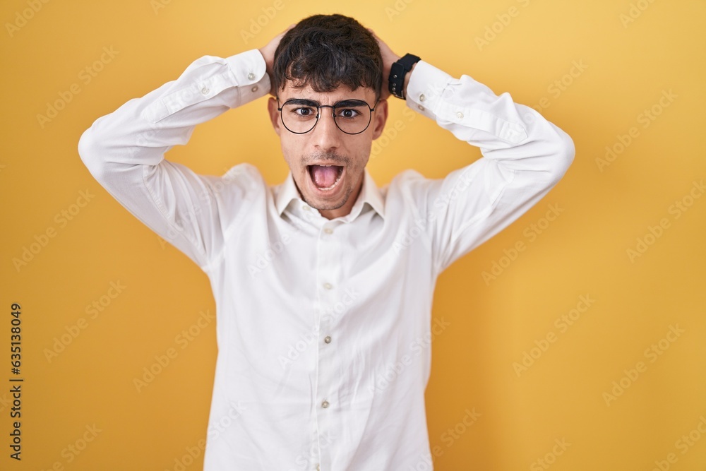 Young hispanic man standing over yellow background crazy and scared with hands on head, afraid and surprised of shock with open mouth Young hispanic man standing over yellow background crazy and scared with hands on head, afraid and surprised of shock with open mouth