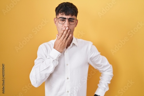 Young hispanic man standing over yellow background bored yawning tired covering mouth with hand. restless and sleepiness.