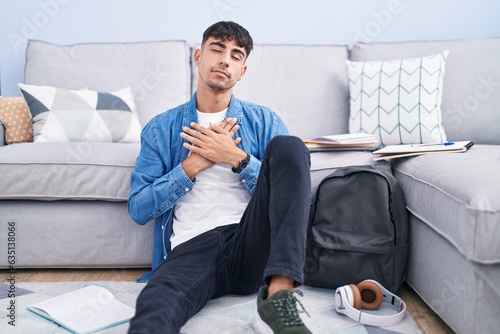Young hispanic man sitting on the floor studying for university smiling with hands on chest with closed eyes and grateful gesture on face. health concept.