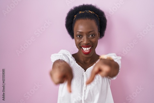 African woman with curly hair standing over pink background pointing to you and the camera with fingers, smiling positive and cheerful