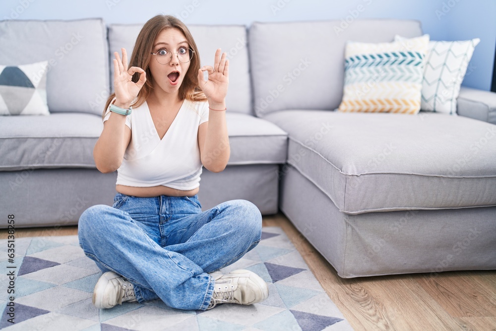 Young caucasian woman sitting on the floor at the living room looking surprised and shocked doing ok approval symbol with fingers. crazy expression Young caucasian woman sitting on the floor at the living room looking surprised and shocked doing ok approval symbol with fingers. crazy expression