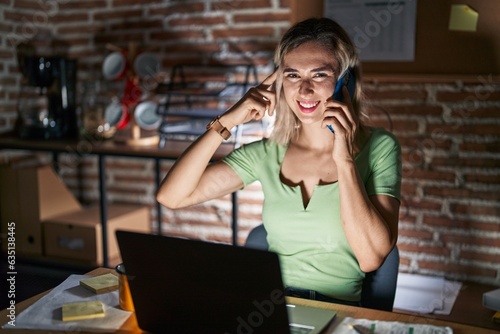 Young beautiful woman working at the office at night speaking on the phone smiling pointing to head with one finger, great idea or thought, good memory
