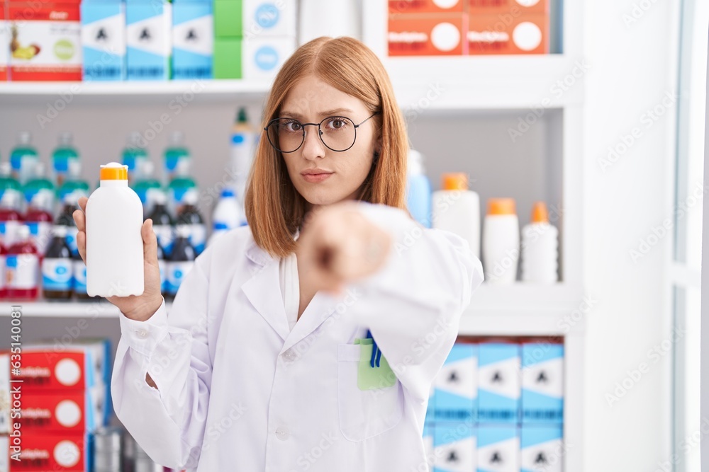 Young redhead woman working at pharmacy drugstore holding sun screen pointing with finger to the camera and to you, confident gesture looking serious