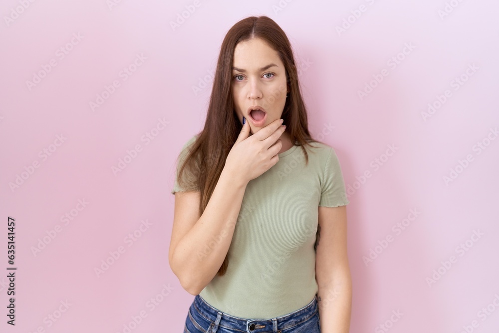 Beautiful brunette woman standing over pink background looking fascinated with disbelief, surprise and amazed expression with hands on chin Beautiful brunette woman standing over pink background looking fascinated with disbelief, surprise and amazed expression with hands on chin