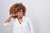 Young hispanic woman with curly hair standing over white background smiling with hand over ear listening an hearing to rumor or gossip. deafness concept.