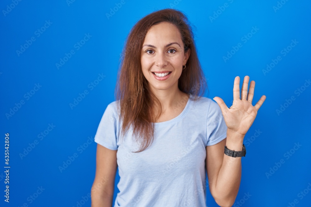 Fototapeta premium Brunette woman standing over blue background showing and pointing up with fingers number five while smiling confident and happy.