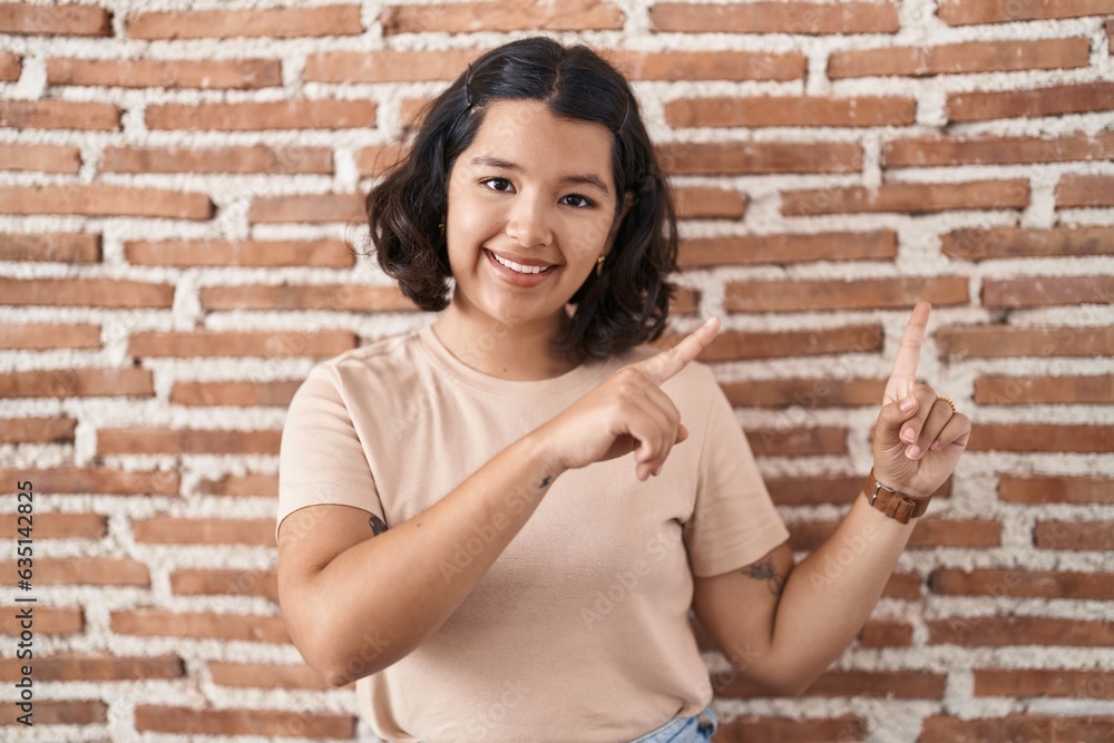 Young hispanic woman standing over bricks wall smiling and looking at the camera pointing with two hands and fingers to the side. Young hispanic woman standing over bricks wall smiling and looking at the camera pointing with two hands and fingers to the side.