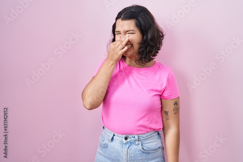 Young hispanic woman standing over pink background smelling something stinky and disgusting, intolerable smell, holding breath with fingers on nose. bad smell photo