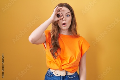 Caucasian woman standing over yellow background doing ok gesture shocked with surprised face, eye looking through fingers. unbelieving expression.