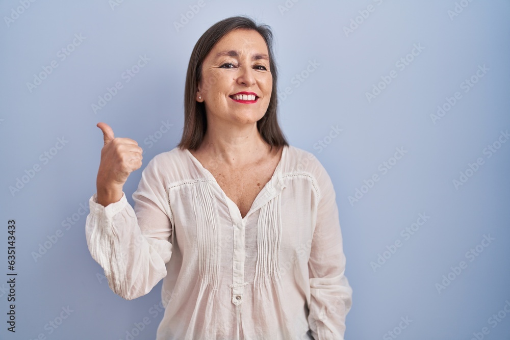 Middle age hispanic woman standing over blue background doing happy thumbs up gesture with hand. approving expression looking at the camera showing success. Middle age hispanic woman standing over blue background doing happy thumbs up gesture with hand. approving expression looking at the camera showing success.
