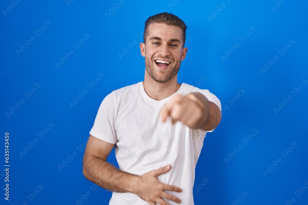 Young caucasian man standing over blue background laughing at you, pointing finger to the camera with hand over body, shame expression Young caucasian man standing over blue background laughing at you, pointing finger to the camera with hand over body, shame expression