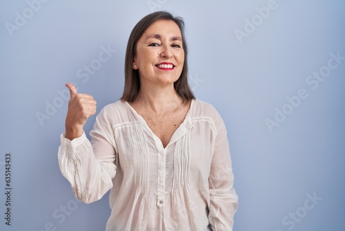 Middle age hispanic woman standing over blue background doing happy thumbs up gesture with hand. approving expression looking at the camera showing success.
