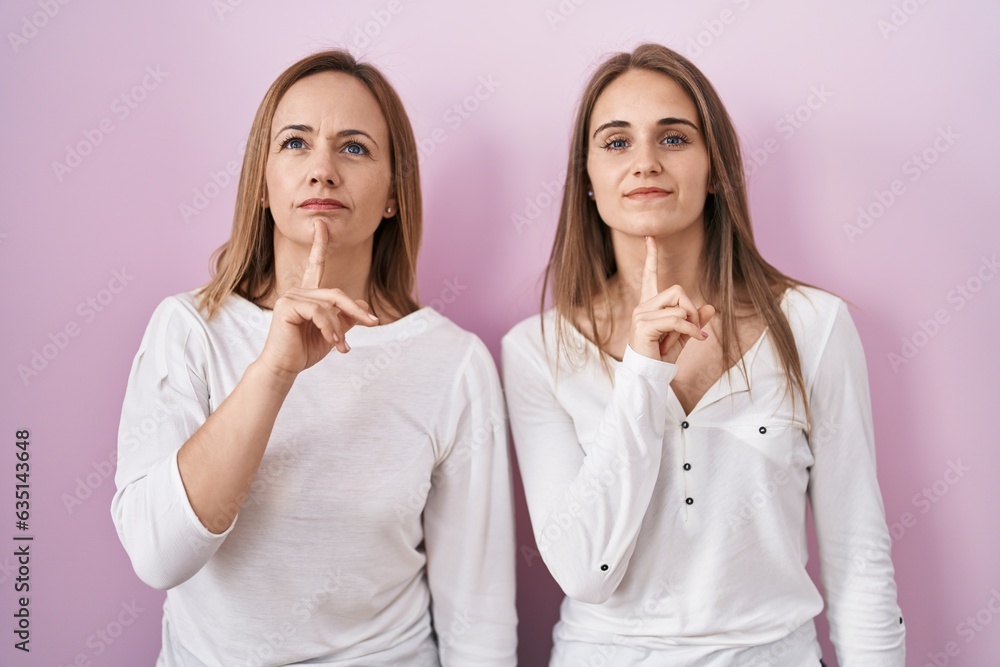Middle age mother and young daughter standing over pink background thinking concentrated about doubt with finger on chin and looking up wondering Middle age mother and young daughter standing over pink background thinking concentrated about doubt with finger on chin and looking up wondering