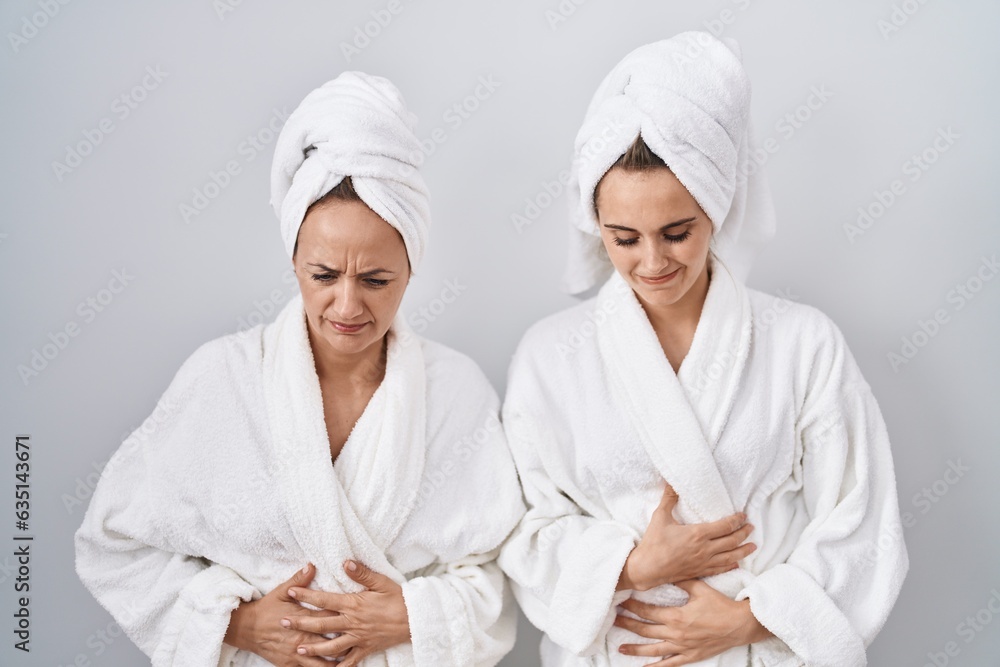 Middle age woman and daughter wearing white bathrobe and towel with hand on stomach because nausea, painful disease feeling unwell. ache concept. Middle age woman and daughter wearing white bathrobe and towel with hand on stomach because nausea, painful disease feeling unwell. ache concept.