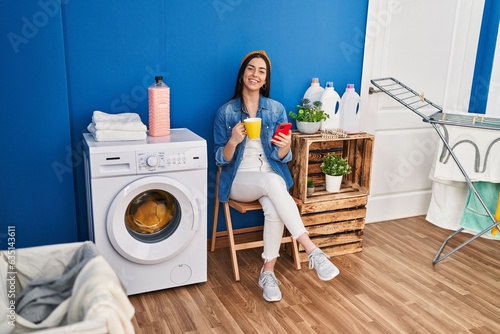 Young beautiful hispanic woman using smartphone drinking coffee waiting for washing machine at laundry room
