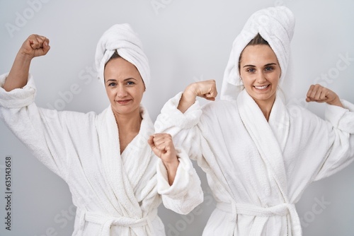 Middle age woman and daughter wearing white bathrobe and towel showing arms muscles smiling proud. fitness concept.