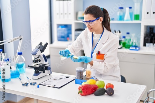 Foto Young beautiful hispanic woman scientist pouring liquid on piece of apple at lab