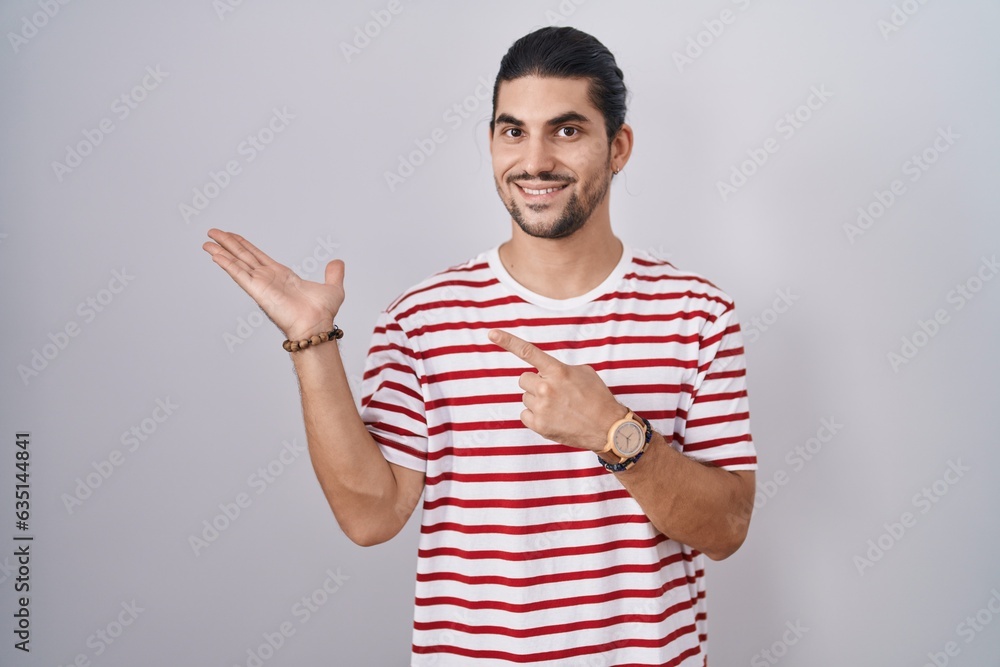 Hispanic man with long hair standing over isolated background amazed and smiling to the camera while presenting with hand and pointing with finger. Hispanic man with long hair standing over isolated background amazed and smiling to the camera while presenting with hand and pointing with finger.