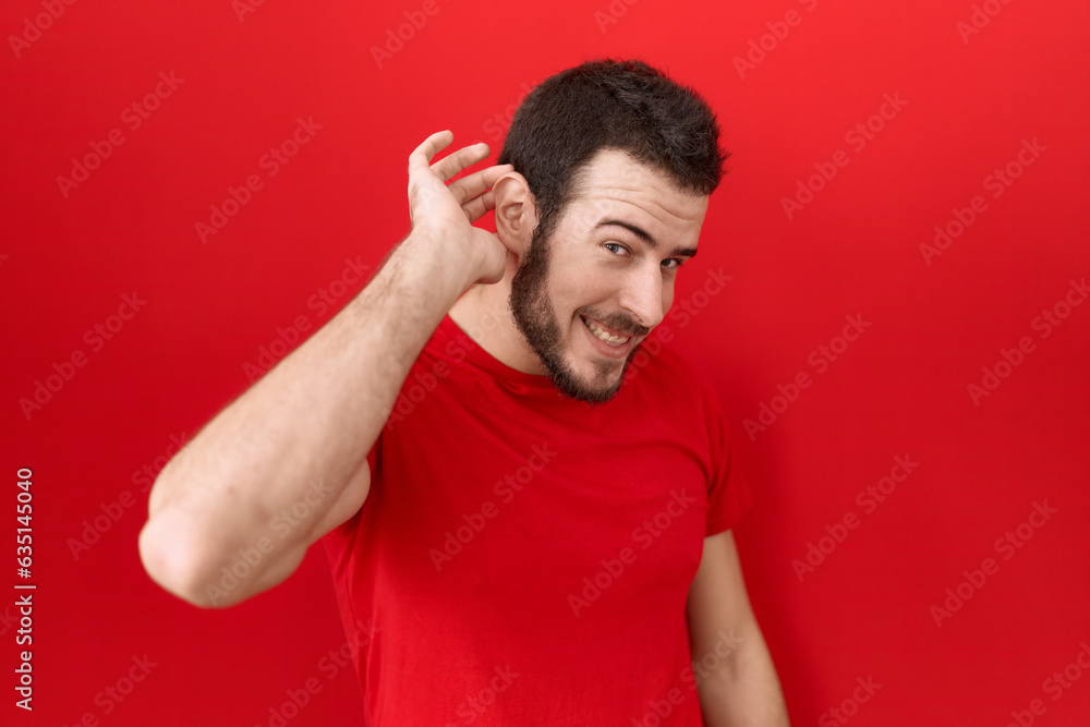 Young hispanic man wearing casual red t shirt smiling with hand over ear listening an hearing to rumor or gossip. deafness concept. Young hispanic man wearing casual red t shirt smiling with hand over ear listening an hearing to rumor or gossip. deafness concept.