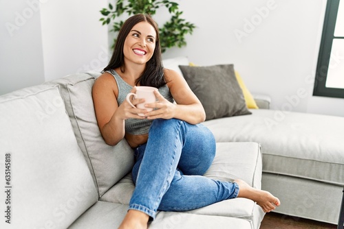 Young beautiful hispanic woman drinking coffee sitting on sofa at home