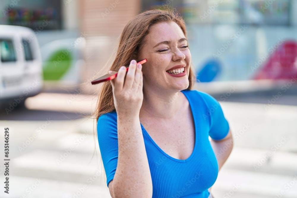 Young redhead woman miling confident listening audio message by the smartphone at street
