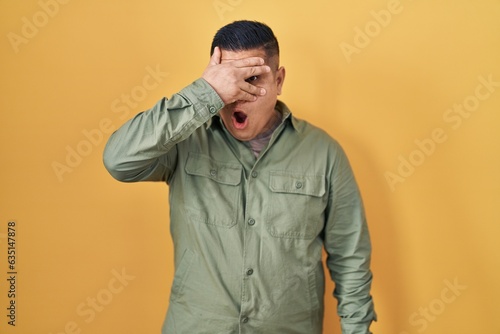 Hispanic young man standing over yellow background peeking in shock covering face and eyes with hand, looking through fingers with embarrassed expression.