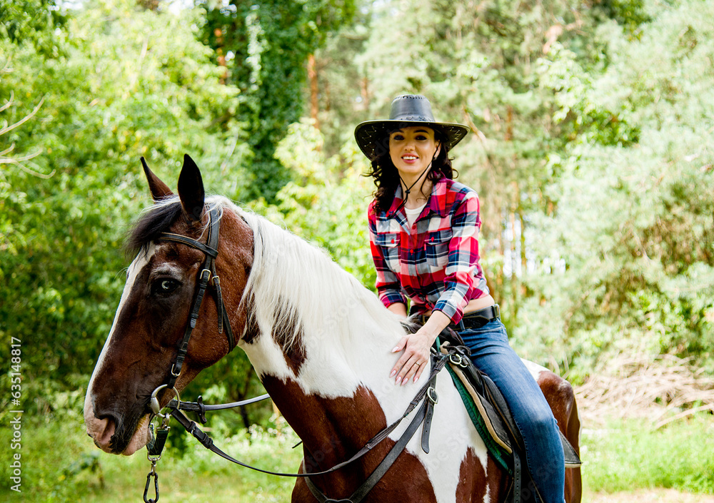 beautiful smiling brunette cowboy woman in hat and red checkered shirt riding horse in green forest beautiful smiling brunette cowboy woman in hat and red checkered shirt riding horse in green forest