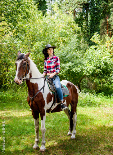 beautiful smiling brunette cowboy woman in hat and red checkered shirt riding horse in green forest 