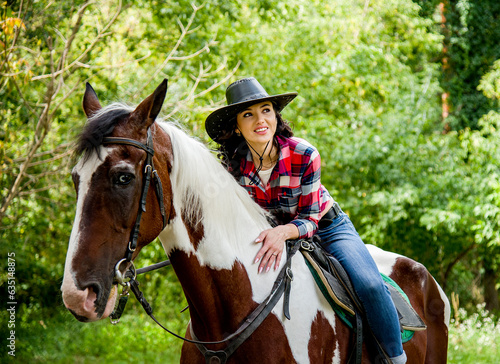 beautiful smiling brunette cowboy woman in hat and red checkered shirt riding and hugging horse in green forest 