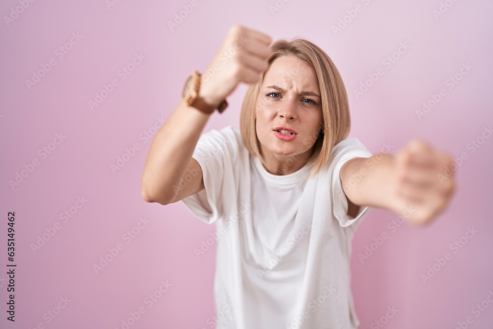 Young caucasian woman standing over pink background angry and mad raising fists frustrated and furious while shouting with anger. rage and aggressive concept. Young caucasian woman standing over pink background angry and mad raising fists frustrated and furious while shouting with anger. rage and aggressive concept.