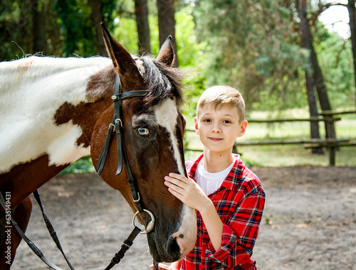 little handsome blonde smiling boy in red checkered shirt hugging horse in green forest on sunny day 