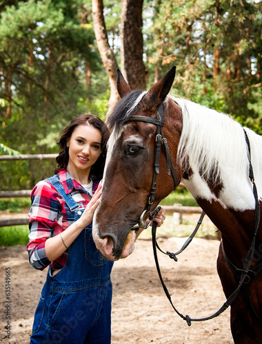 portrait of beautiful smiling brunette cowboy woman in red checkered shirt walking and hugging with horse with blue eyes in green forest on sunny day 