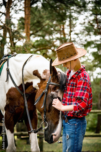 little handsome blonde smiling boy in red checkered shirt and cowboy hat hugging horse in green forest on sunny day 